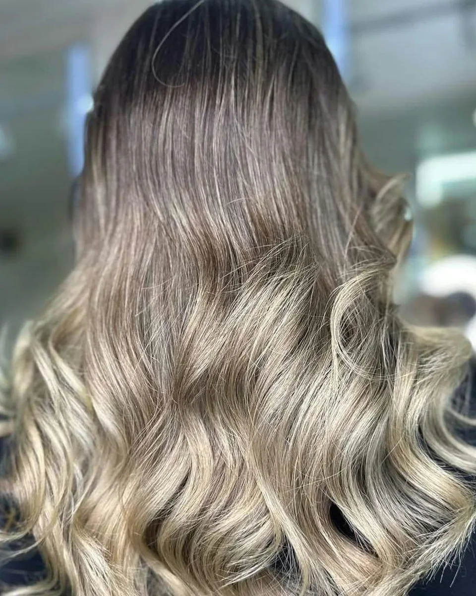 Long, glossy hair with dark roots melting into creamy blonde balayage, styled in soft waves